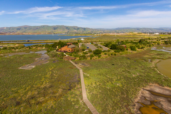 An aerial view of the Environmental Education Center (a large brown angular building) in the center of green and blue wetlands. Large hills are in the distance. Photo credit: Cris Benton, 2013.