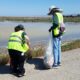 Two volunteers in safety vests pick up litter from the edge of the marsh.